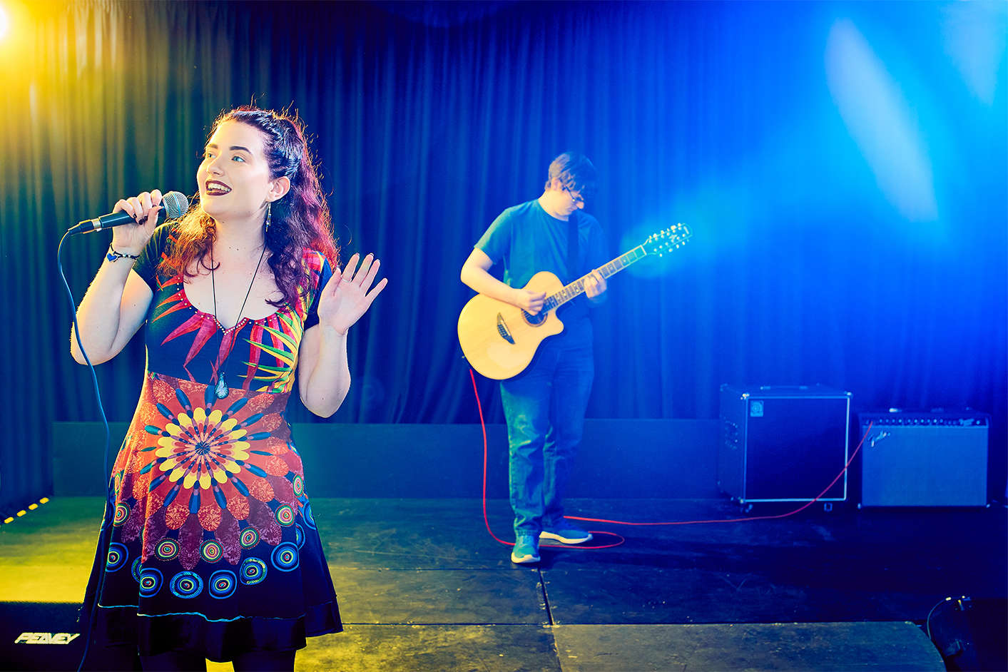 A young female stood on stage singing with a microphone and guitarist playing behind her