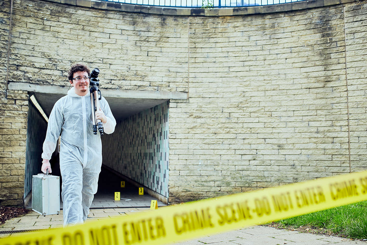 a boy wearing white forensic overalls walking out of a crime scene with a digital camera over his shoulder and metal briefcase