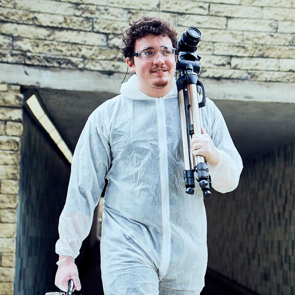 a boy wearing white forensic overalls walking out of a crime scene with a digital camera over his shoulder and metal briefcase