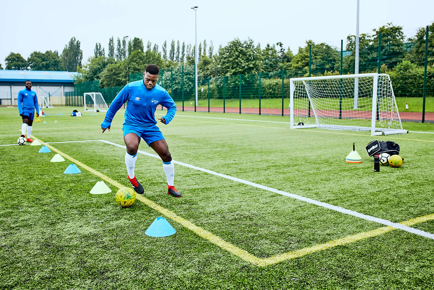 A young black student practising his football skills dribbling around cones on a 3G astroturf pitch