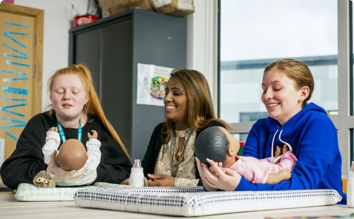 Two female students in a childcare class with their tutor and practise dolls
