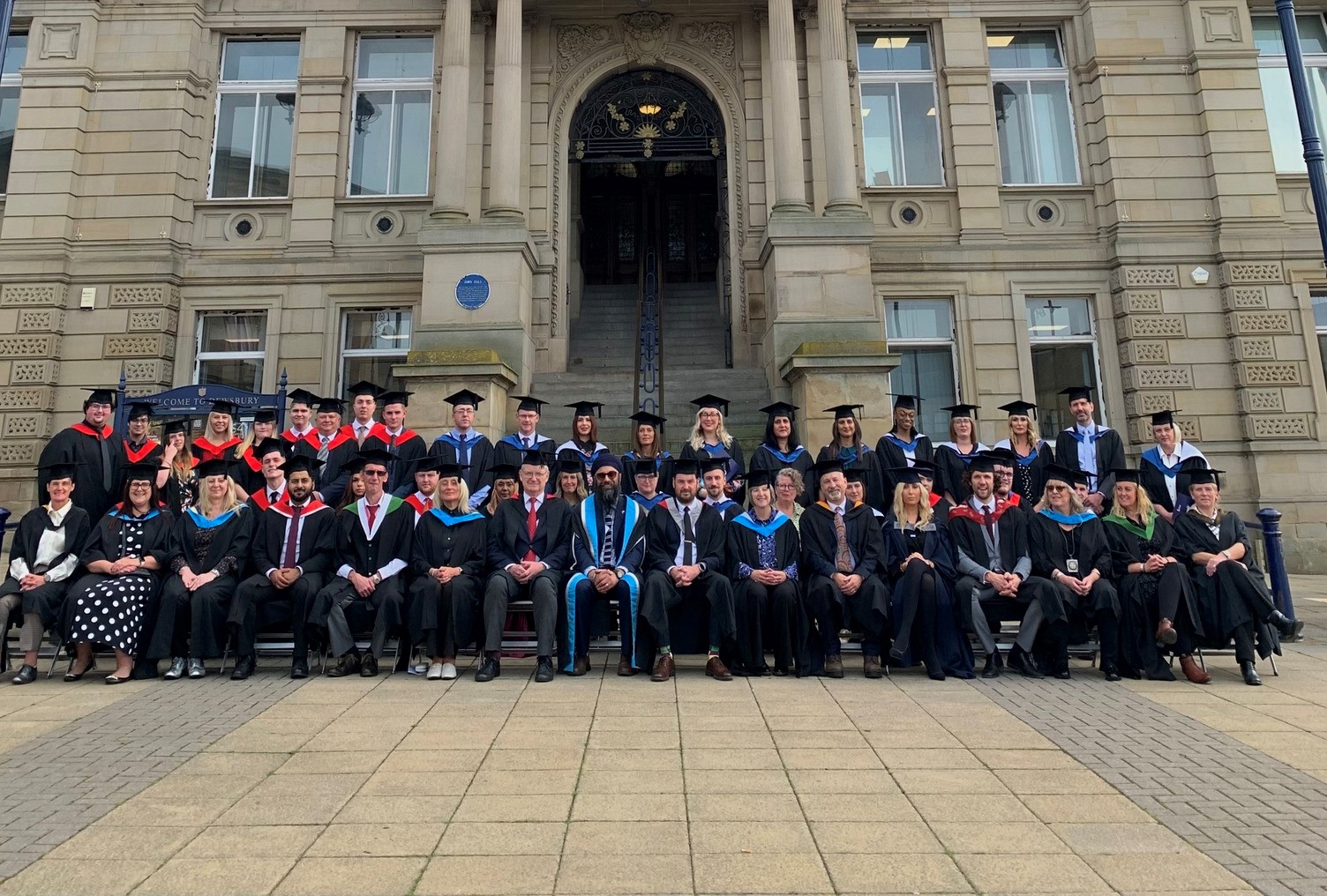 Students and staff in graduation robes sit outside Dewsbury Town Hall doors in two lines