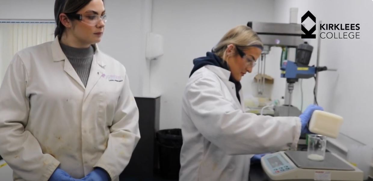 Woman in lab coat stands on left looking at women in lab coat on right using laboratory equipment