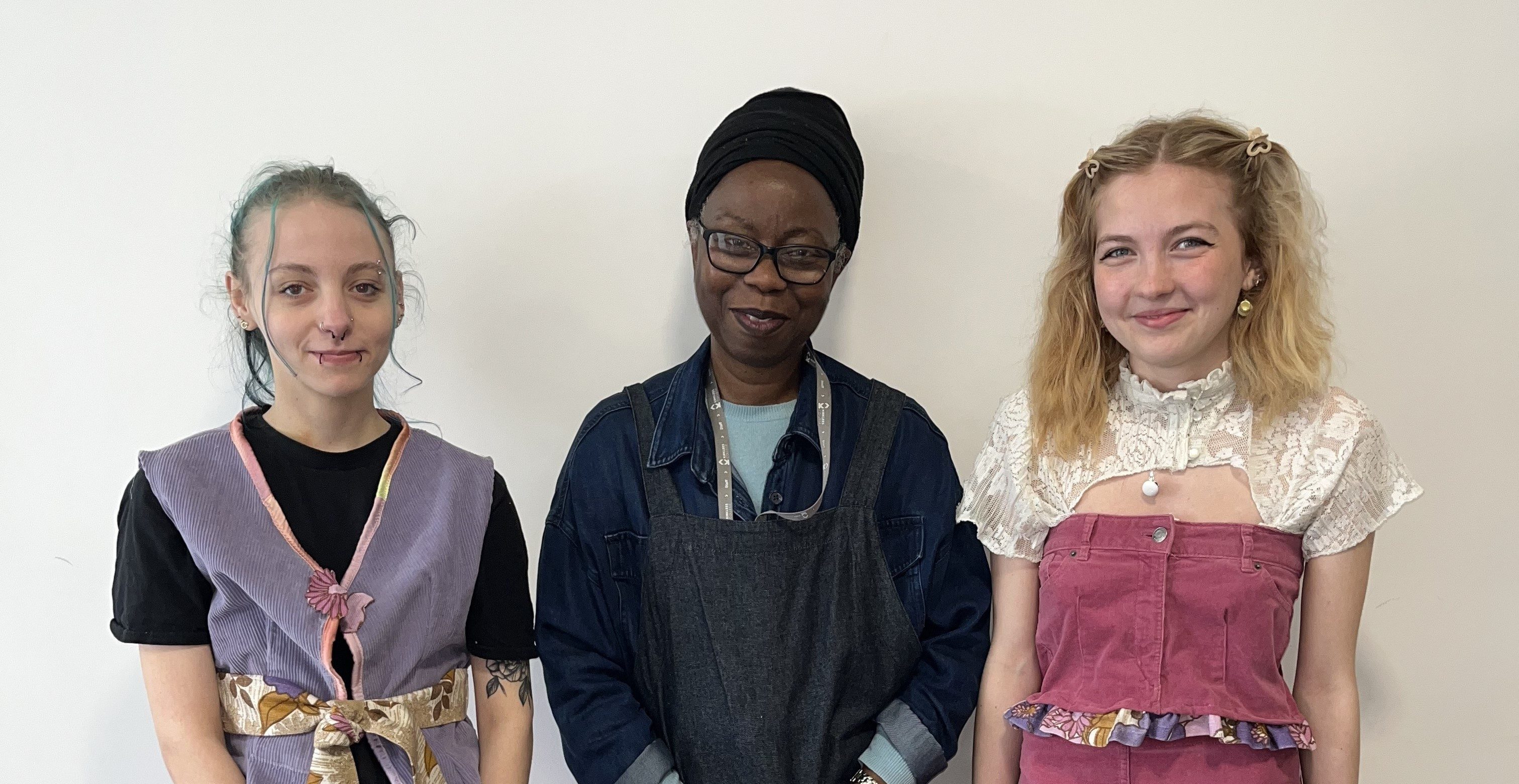 Two students pose for a photo with their tutor, stood in middle, wearing their fashion designs