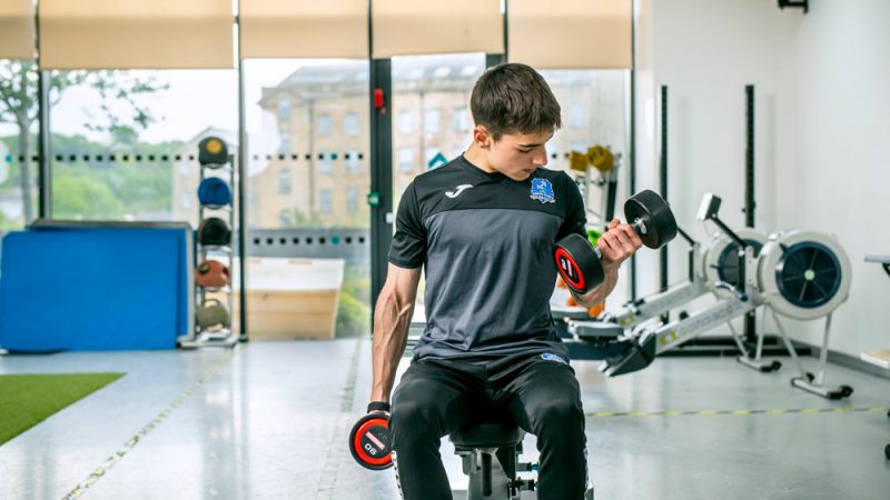Male Sports Student Performing Bicep Curls with Dumbells in Waterfront Gym
