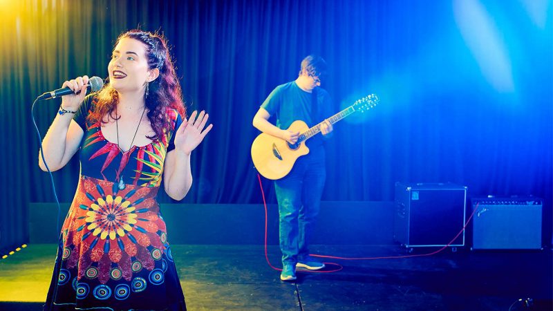 A young female stood on stage singing with a microphone and guitarist playing behind her