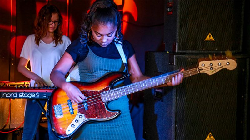 A young black female student playing a guitar on stage in front of a drummer