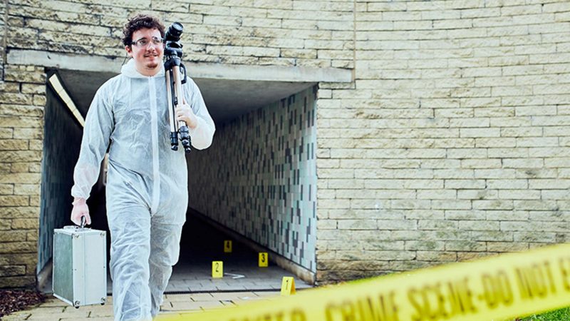 a boy wearing white forensic overalls walking out of a crime scene with a digital camera over his shoulder and metal briefcase